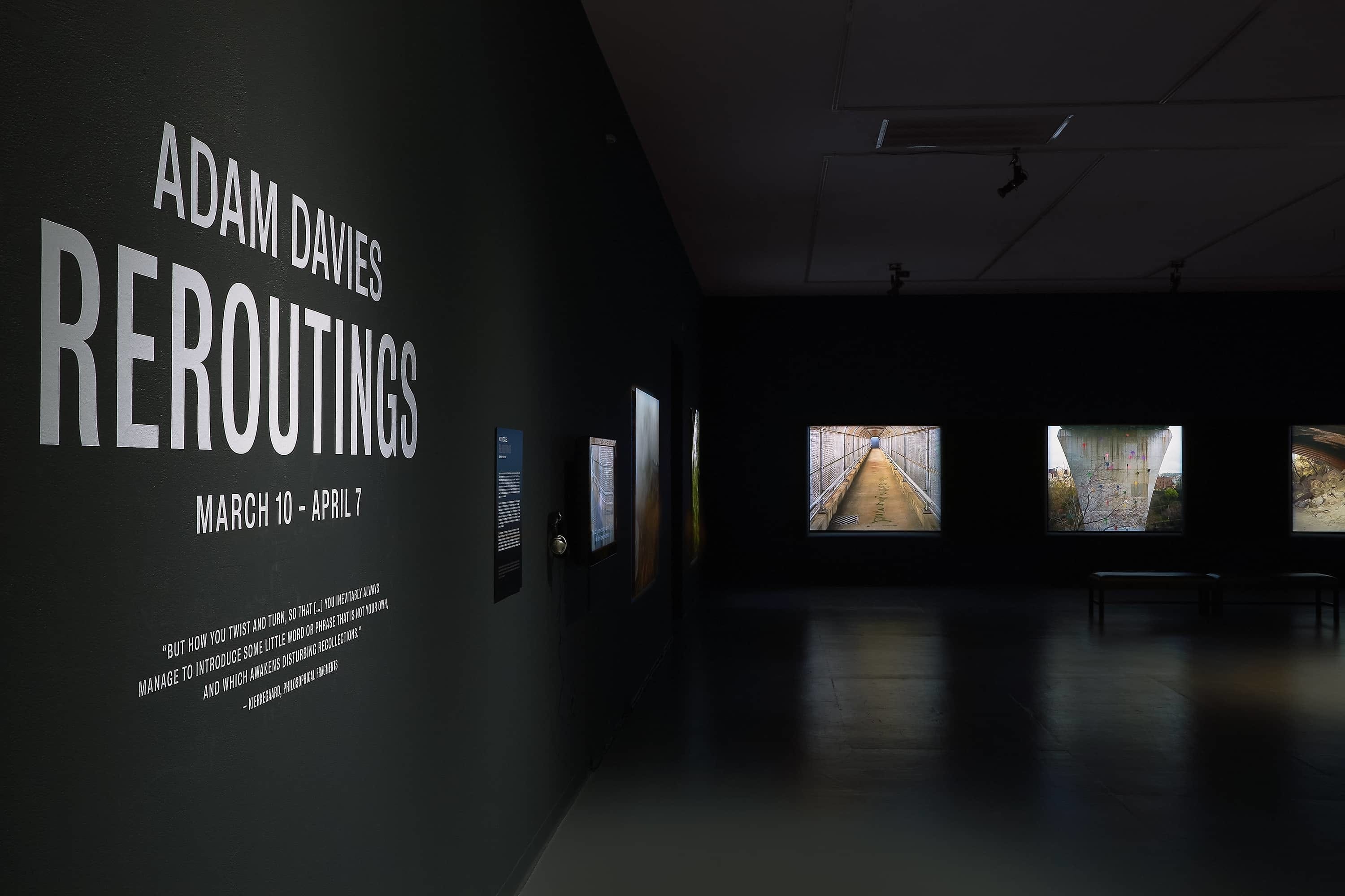 Entrance of exhbition with title text on the wall on left and large photographs illuminated further back in a dark gallery.