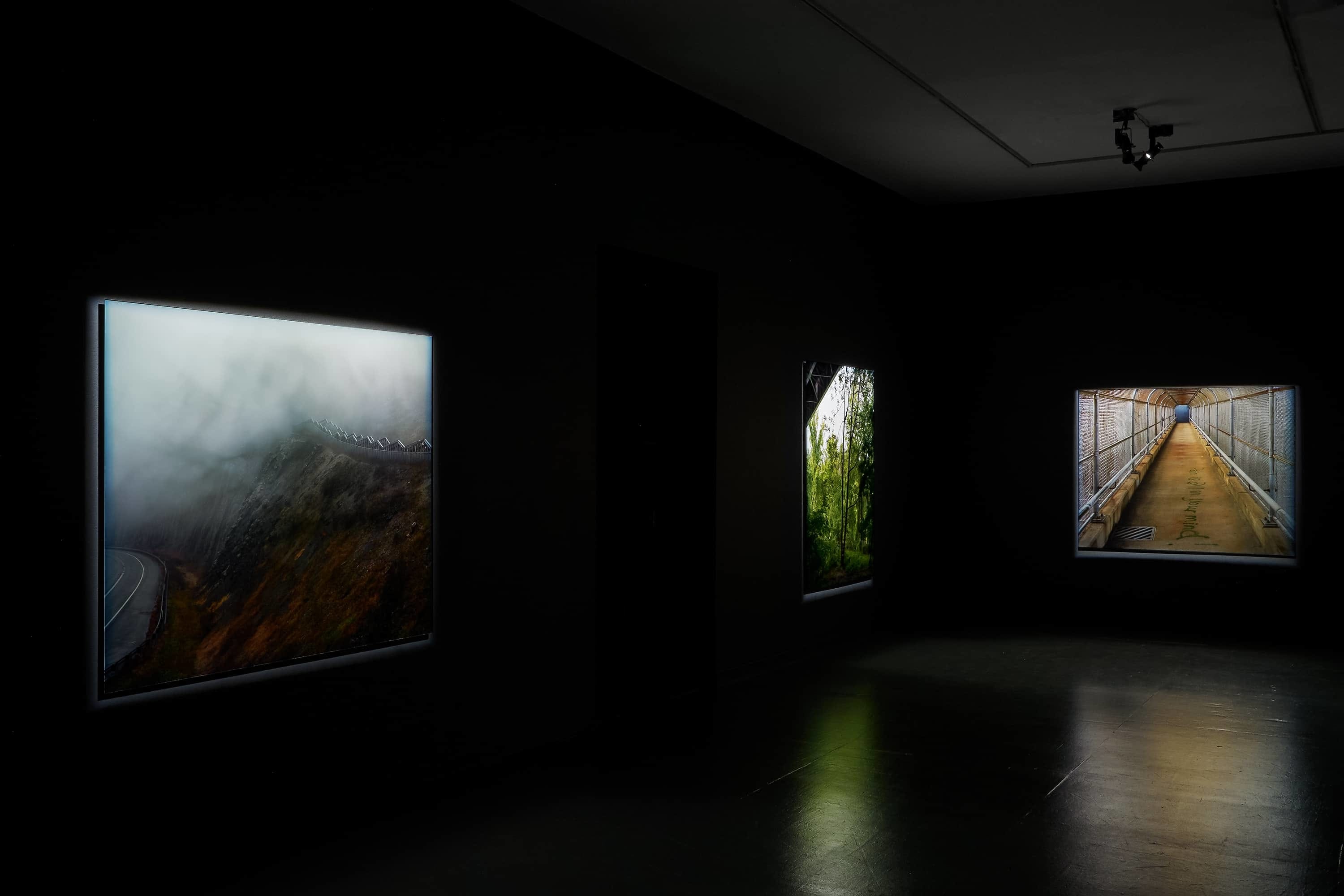 Three large photographs of landscapes and bridges illuminated in a dark gallery, detail.
