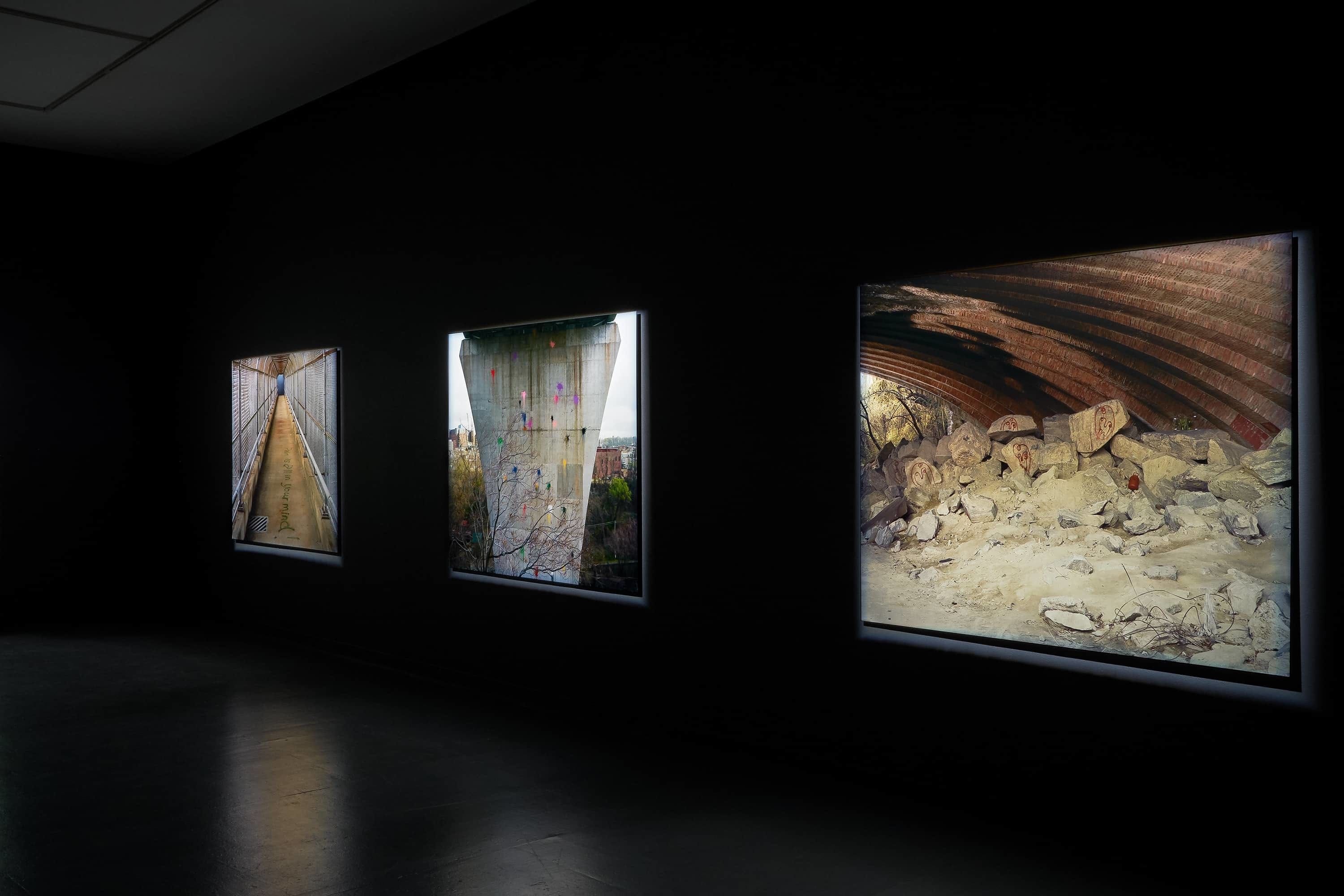 Three large photographs of grafitti on infrastructure illuminated in a dark gallery, detail.