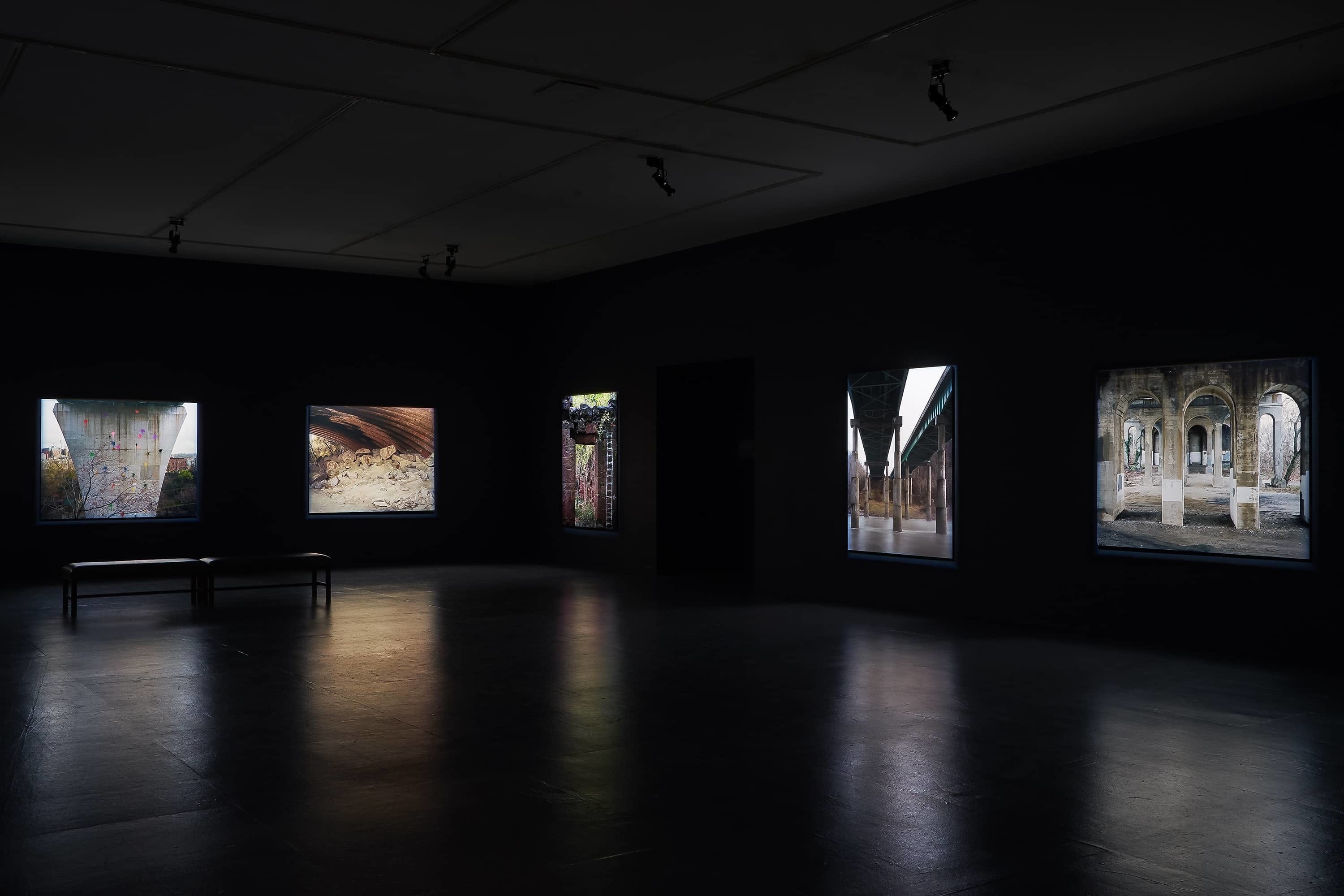 Five large photographs of the substructure of bridges illuminated in a dark gallery, view of two walls.