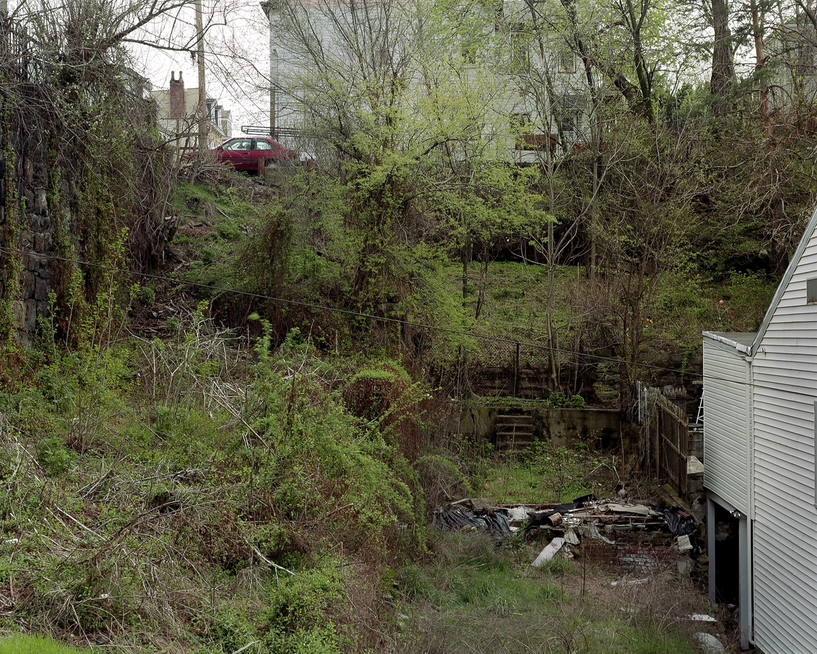 Overgrown and rubble-filled tiered garden in Pittsburgh, on the street above and behind the house a red car is parked.