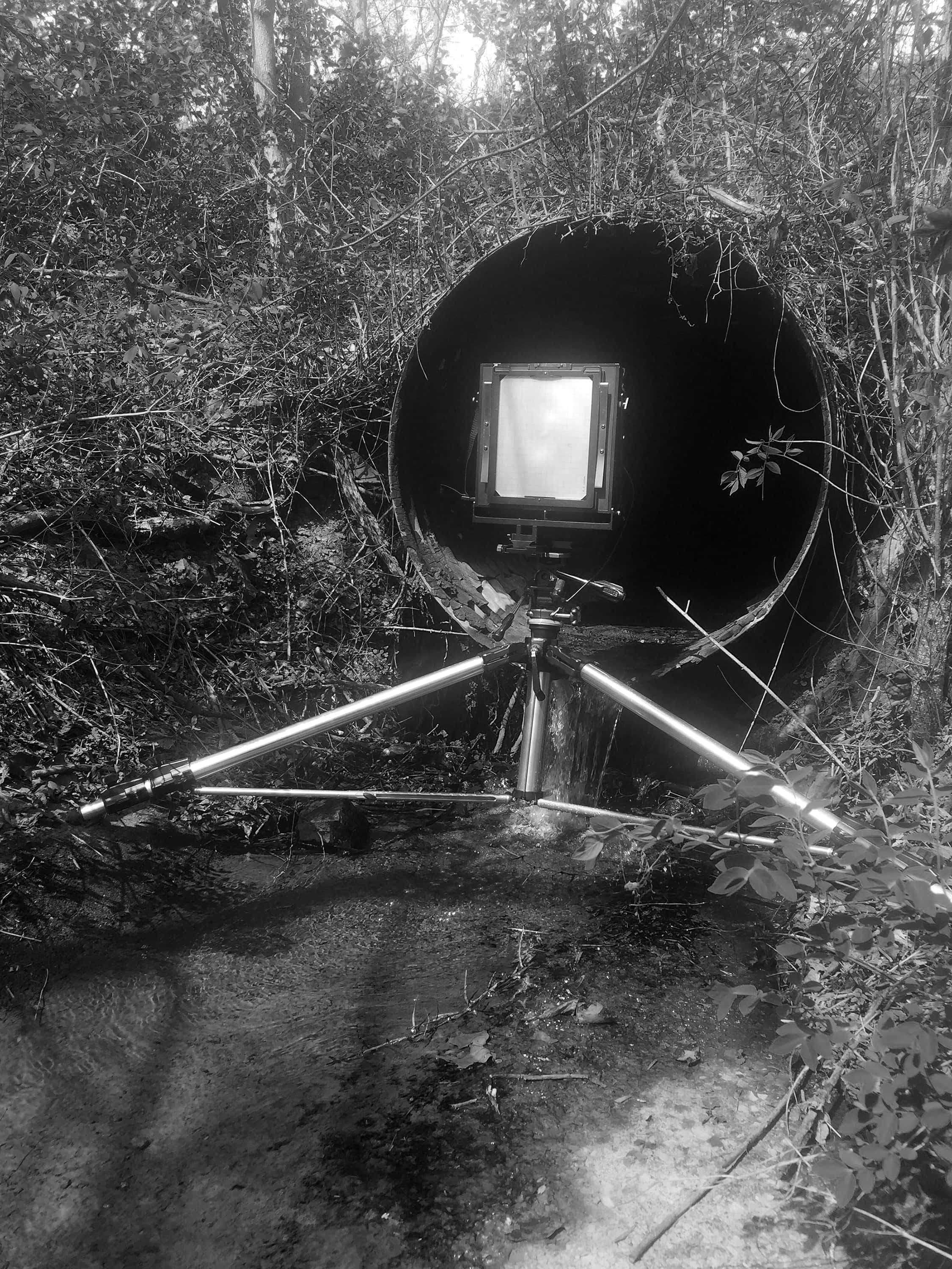 In the woods, a large-format camera on tripod is facing into a large metal culvert out of which a stream flowing.