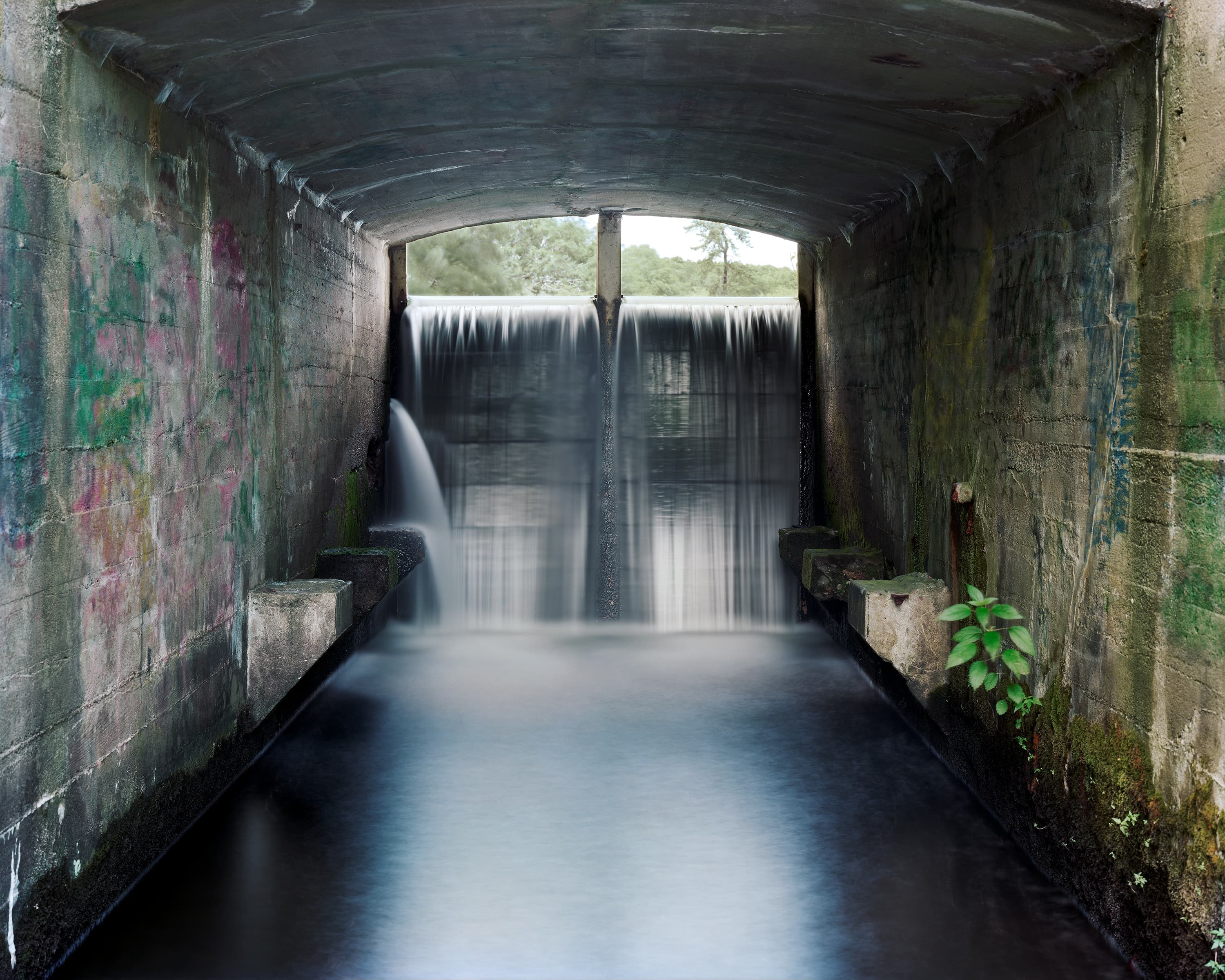 Water from an upper pond pours over a concrete dam at the end of a graffiti lined tunnel.