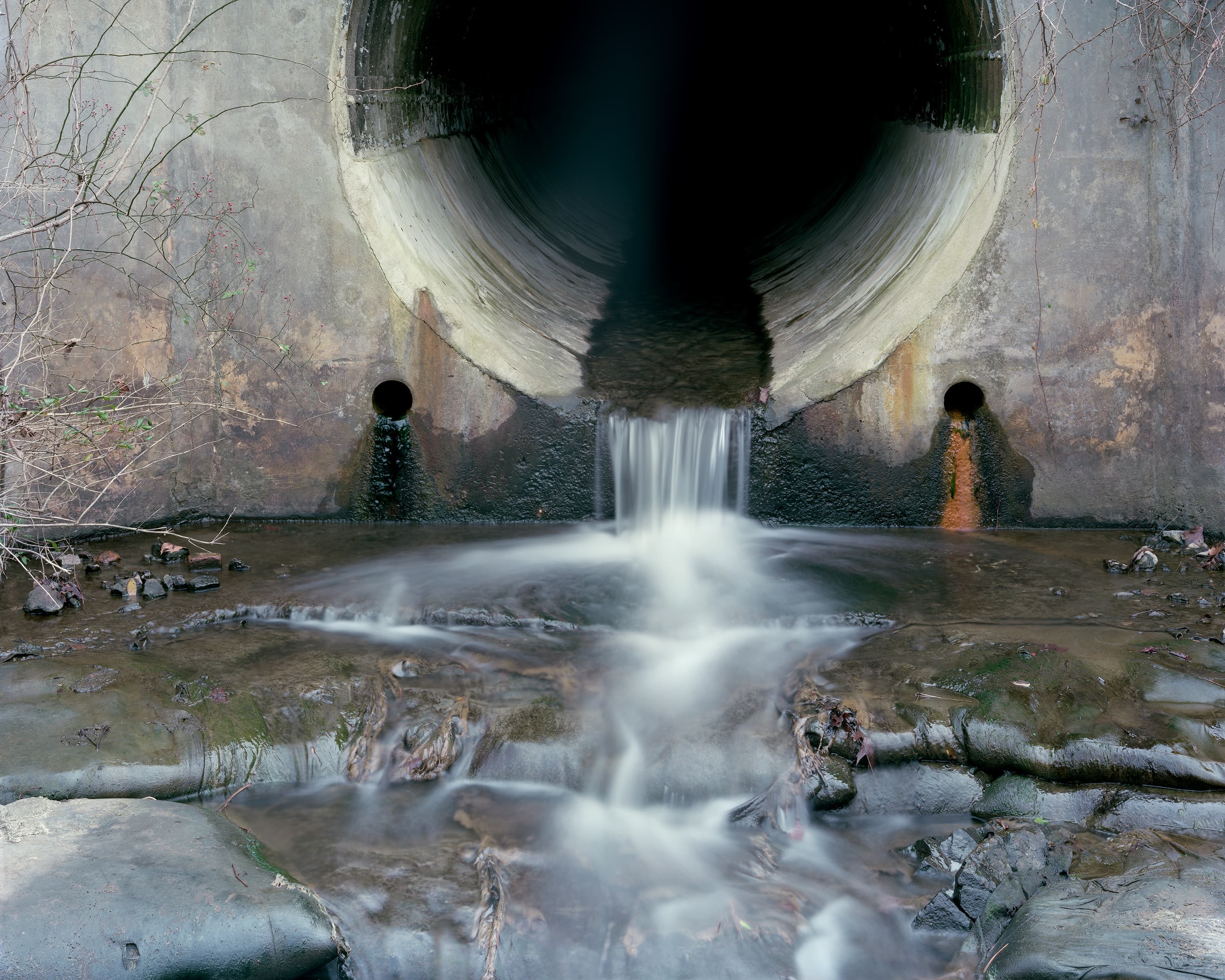 Water runs out of the end of a concrete tunnel cut into a hillside. From two small holes on either side, under the tunnel red clay and green/black oxidized copper drip out.