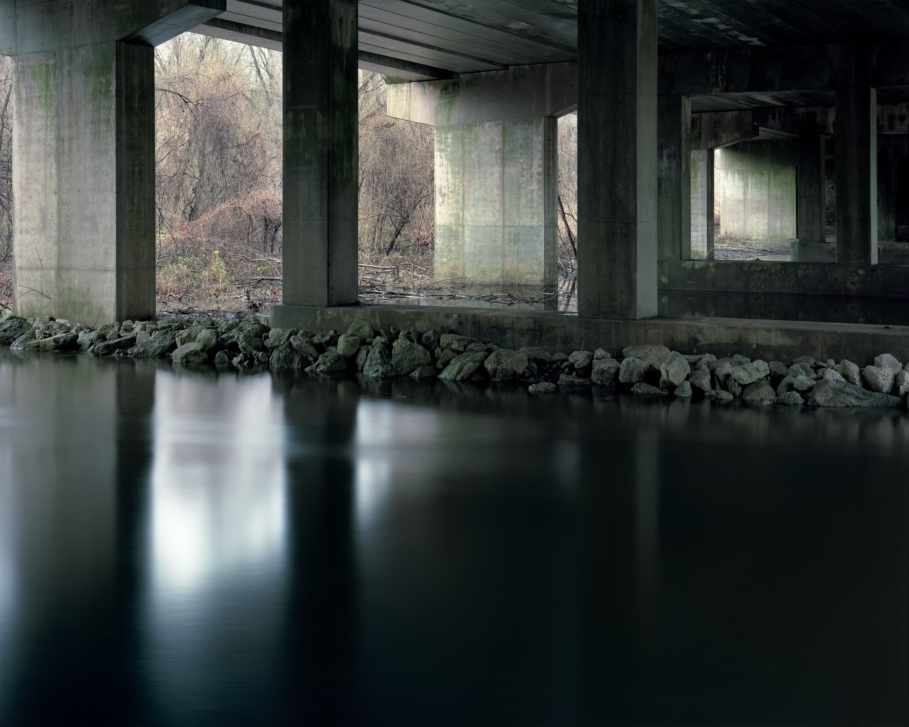 A creek passes under a highway with multiple concrete piers, at one end is an overgrown forest in the fall.