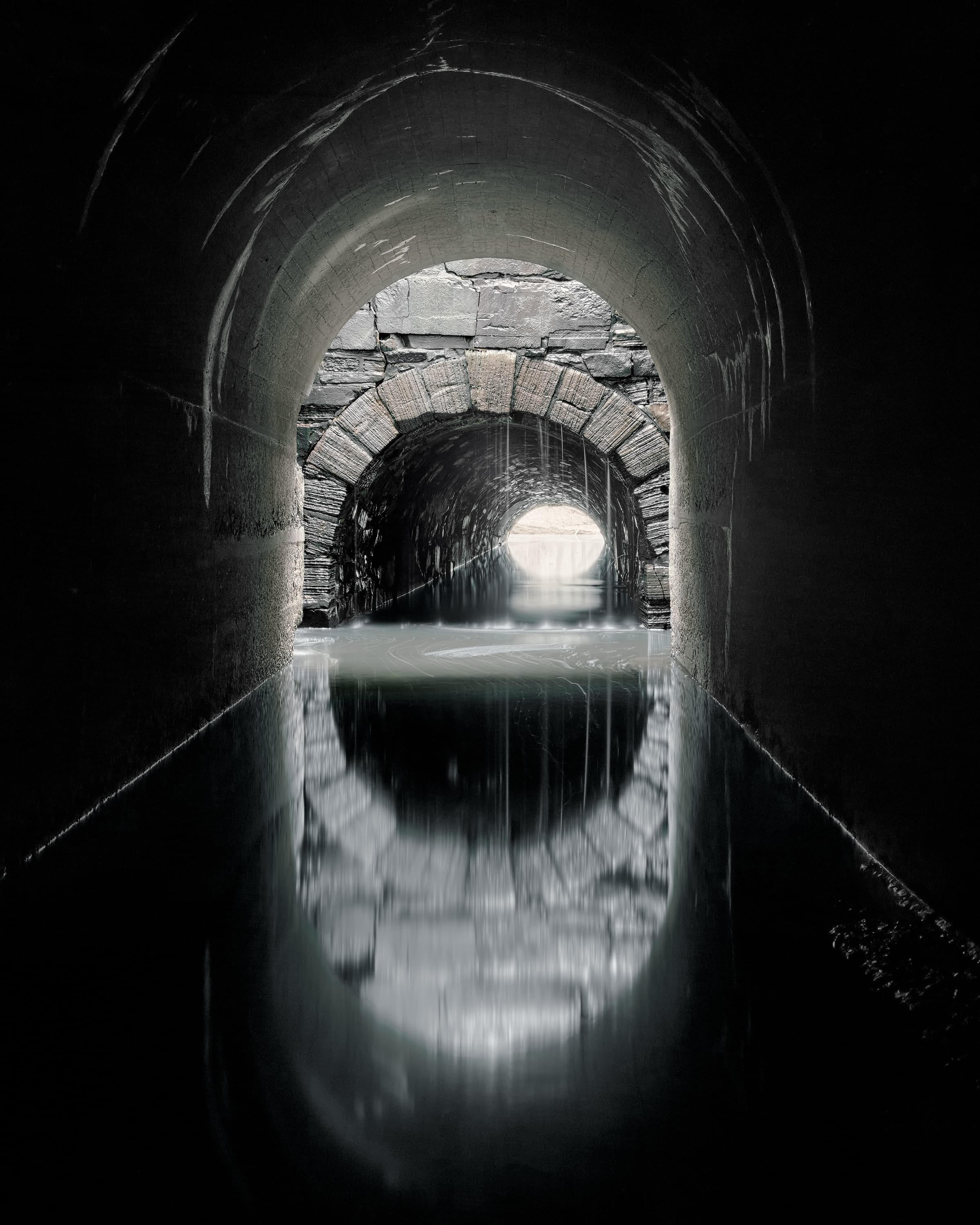 A deep stream runs through two long tunnels, the first cast in concrete, the second in stone. In the second tunnel water is pouring from the roof.