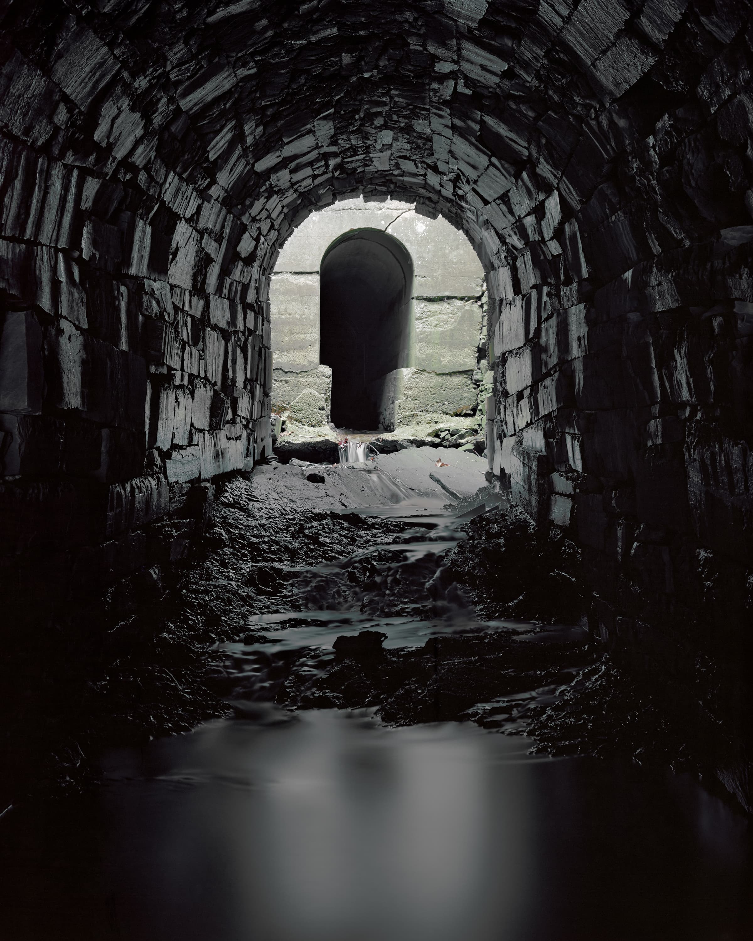 A stream runs through a dark tunnel lined with irregular stones, at the end is a shaft of light and second tunnel cast in concrete, shaped like a keyhole.