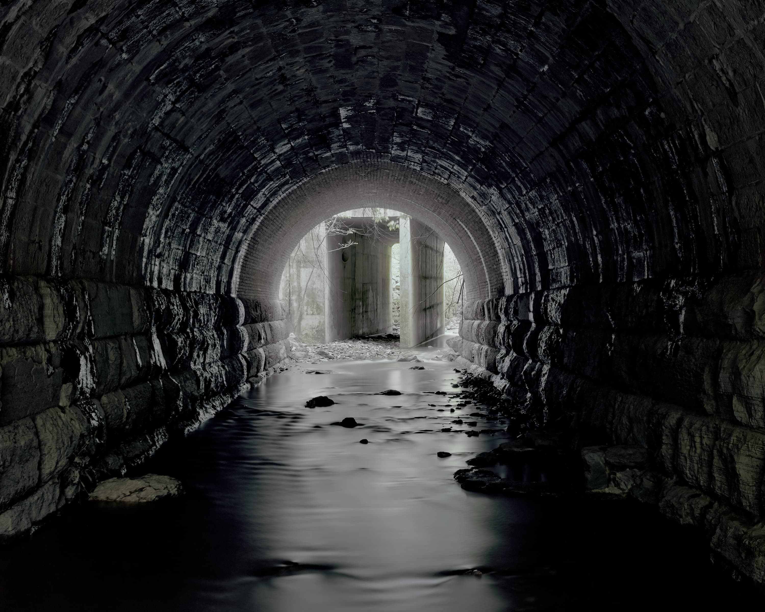 A stream runs through a dark tunnel lined with stones, at the end in the daylight are two abandoned concrete piers.