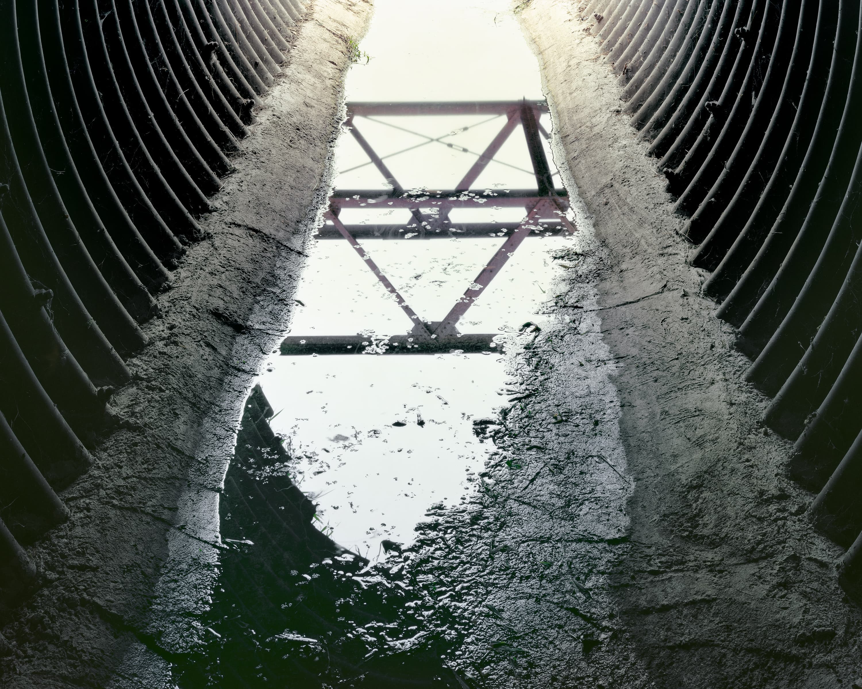 Interior of a metal pipe culvert filled with shallow water, in the reflection in the water the frame of an abandoned bridge is visible.