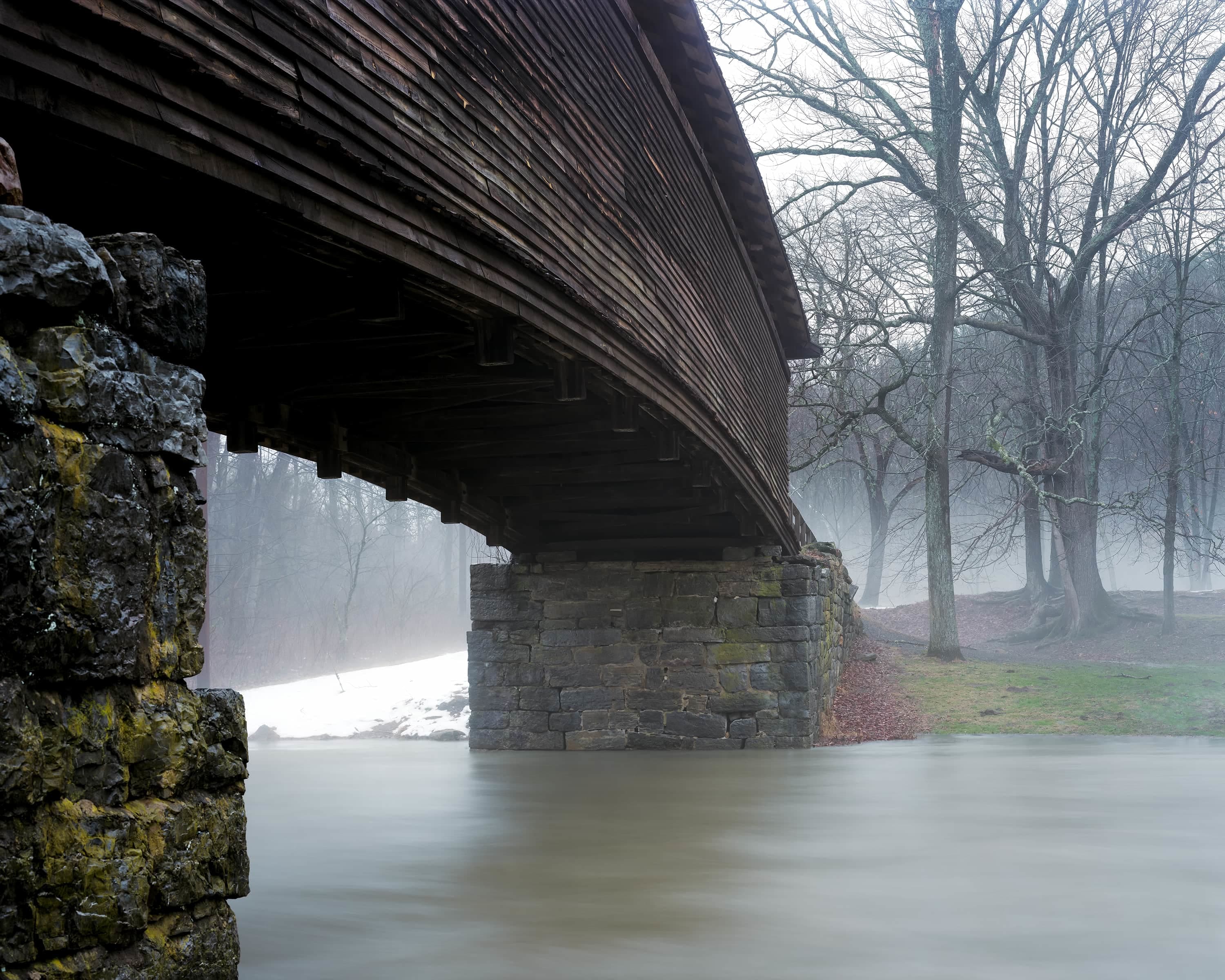 Wooden covered bridge over a small stream in the very early spring, on the left side of the bridge snow is still on the ground, on the right it is all melted.