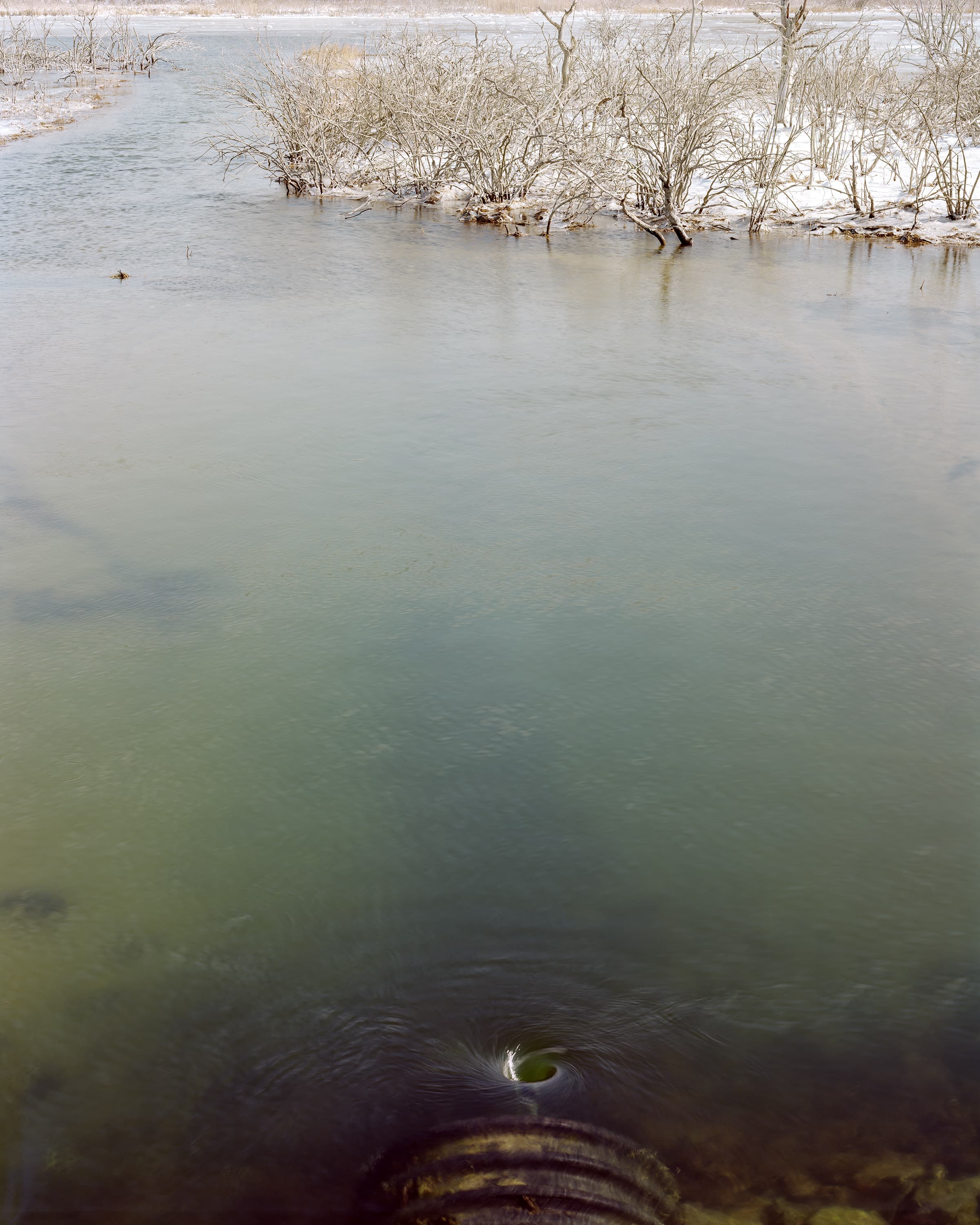 In a pond in the winter, a whirlpool is caused by a large metal pipe in the foreground.