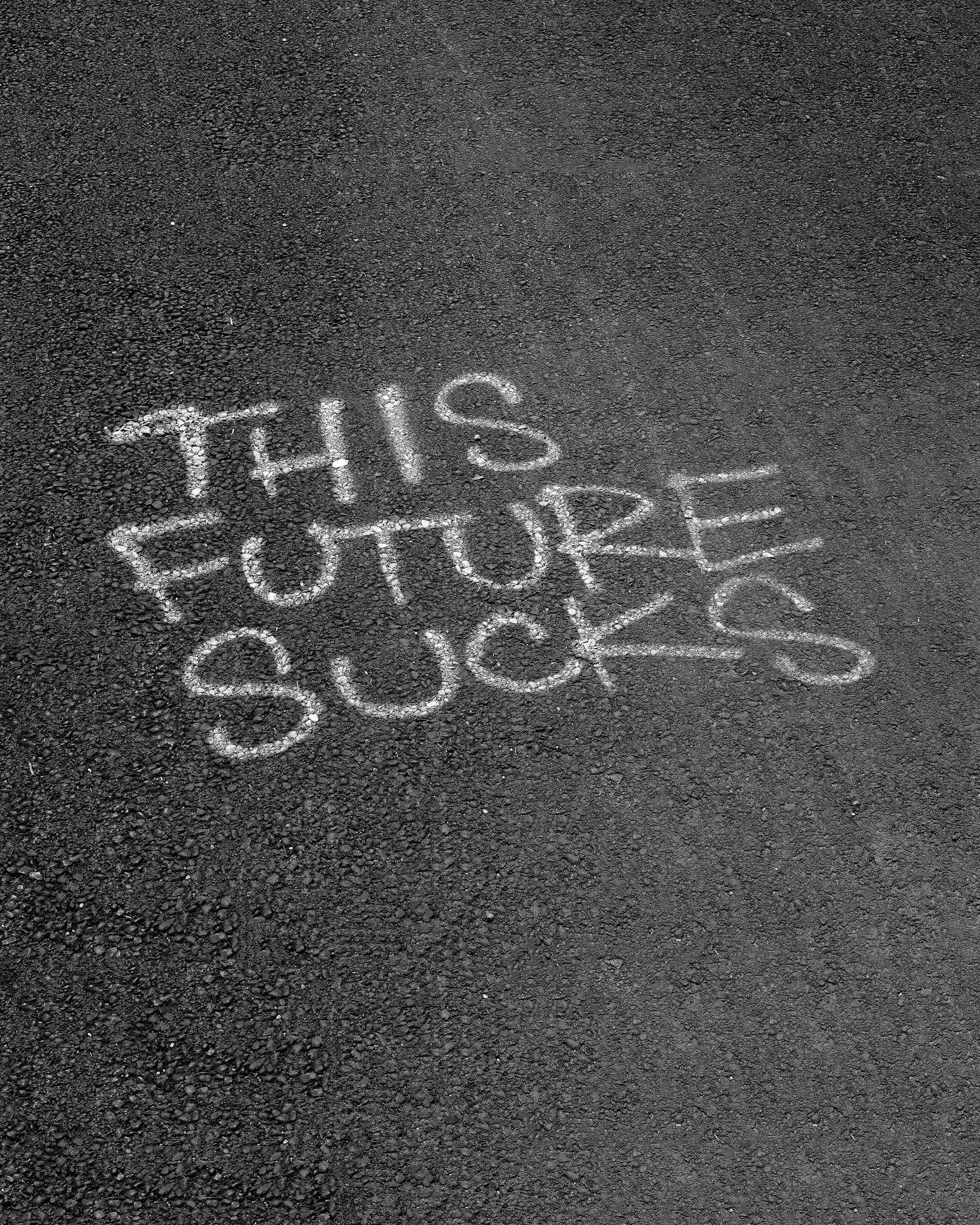 Graffiti with the text, This Future Sucks, written on asphalt surface of a bike path.