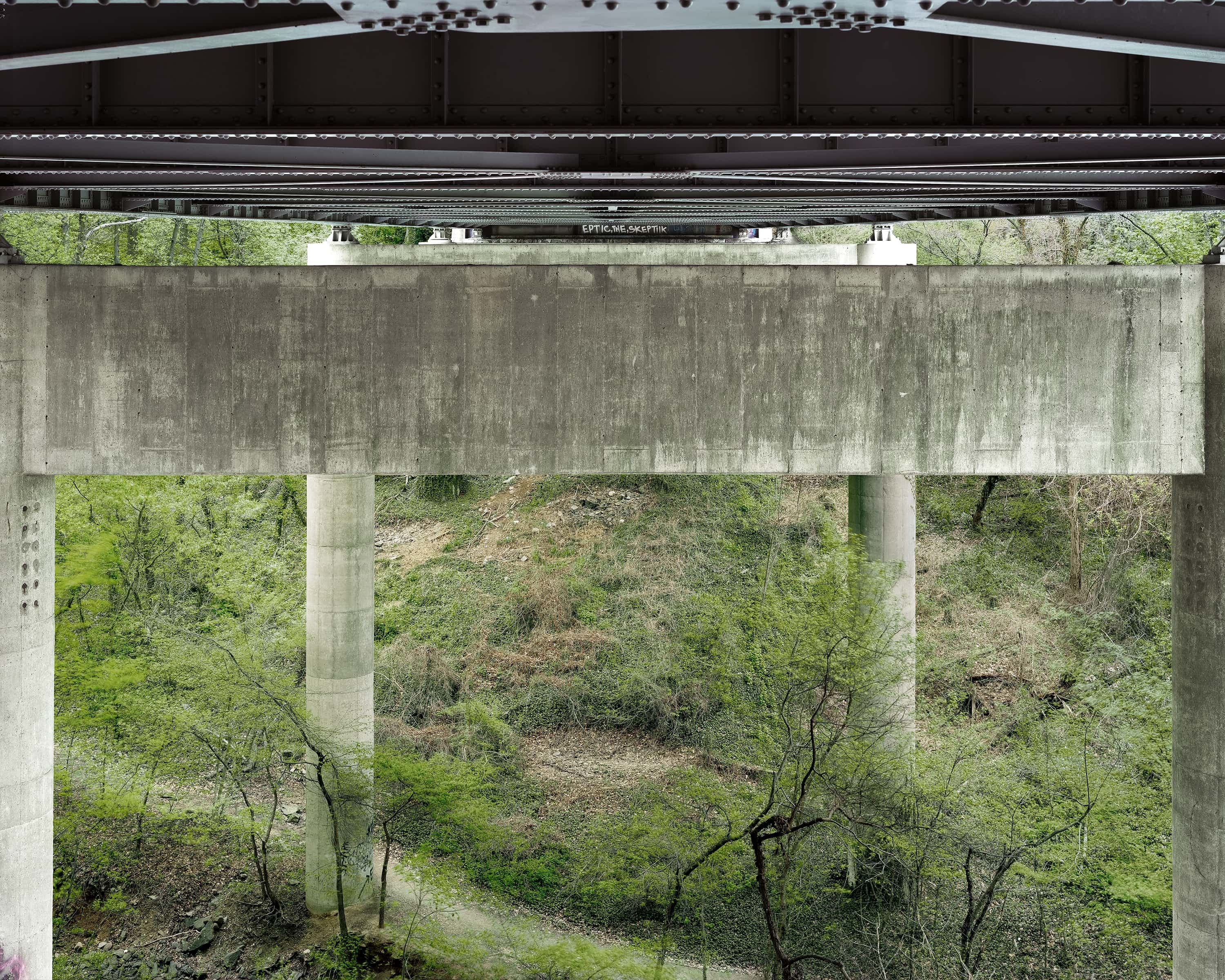 Directly under the deck of a highway looking over the valley below, graffiti in painted on top of a pier.